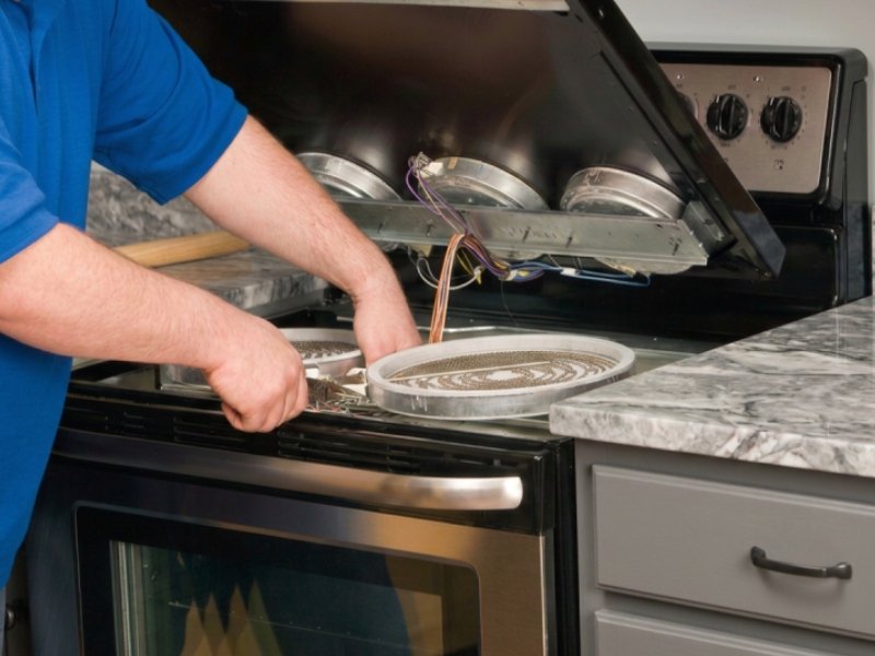 A range repair technician works on the kitchen range, wires are visible inside the stove and a control panel at the back.