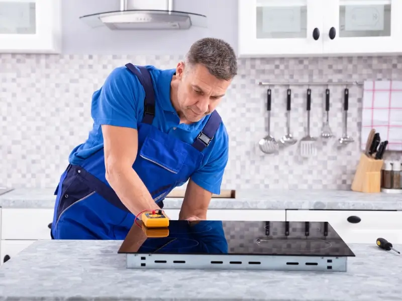 A technician in blue workwear performs cooktop repair in a residential kitchen.