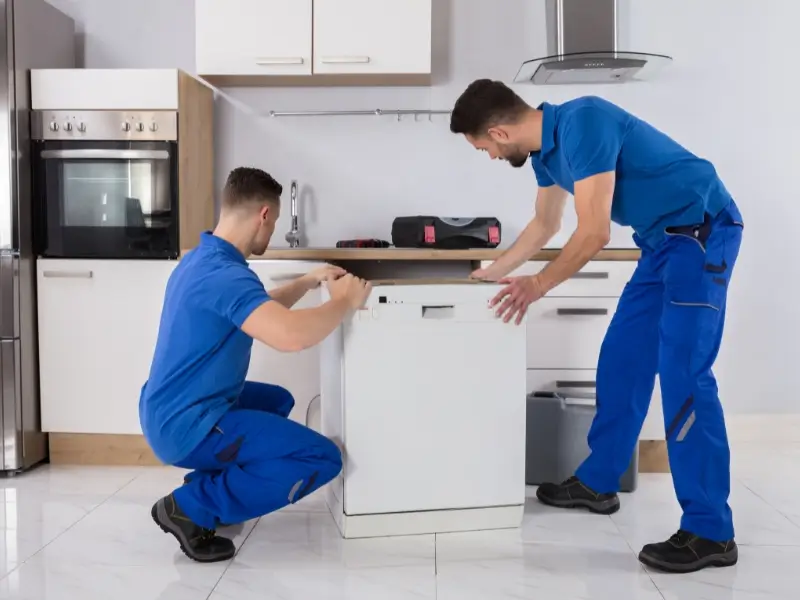 Two technicians in blue uniforms kneel and stand in a kitchen, installing a white dishwasher in a countertop cabinet space - dishwasher installation service.