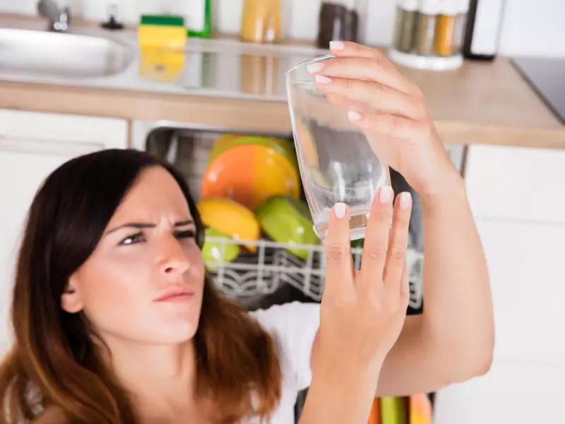 A woman holds up a glass and observes her dishwasher not cleaning well.