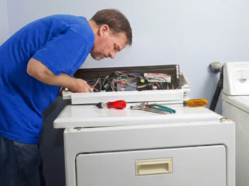 A technician in a blue shirt leans over an open appliance cabinet, doing dryer repair. Wiring components inside while tools and screwdrivers lie on a work surface beside the unit.