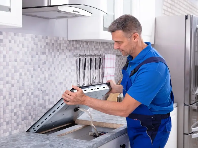 A technician in a blue uniform performs cooktop installation in a modern kitchen, using tools and aligning the unit under the countertop.