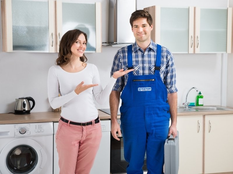An appliance service technician in blue overalls stands in a bright kitchen with an open washing machine door, while another person in a white long-sleeve top and pink pants gestures toward him.
