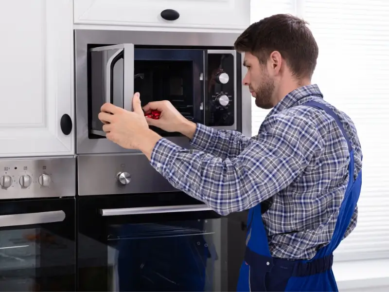 A microwave repair technician in a blue-and-white plaid shirt adjusts a door on a built-in stainless-steel microwave.