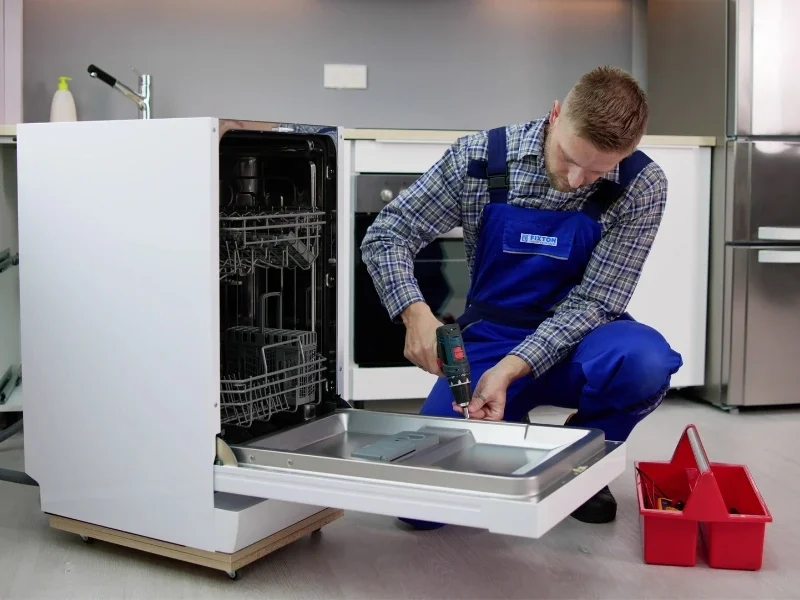Fixton technician in blue overalls kneels in a kitchen, doing panel-ready dishwasher installation with the door open, while red plastic toolbox sits nearby on the floor.