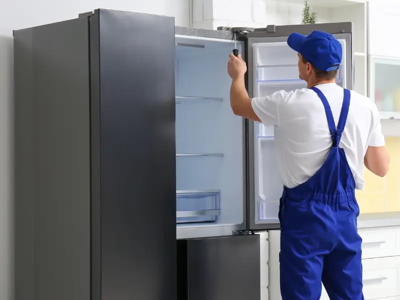 A technician in blue uniform and cap adjusts a refrigerator door during refrigerator installation inside a bright kitchen, using a hand tool.