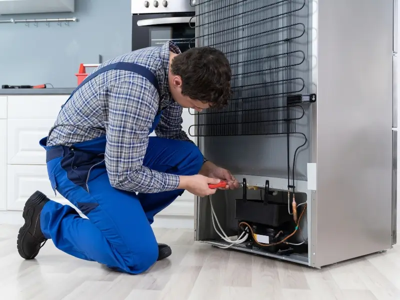 Refrigerator repair technician in blue workwear crouches beside a refrigerator, kneeling on the floor and reaching into the open lower compartment where wiring and components are visible.