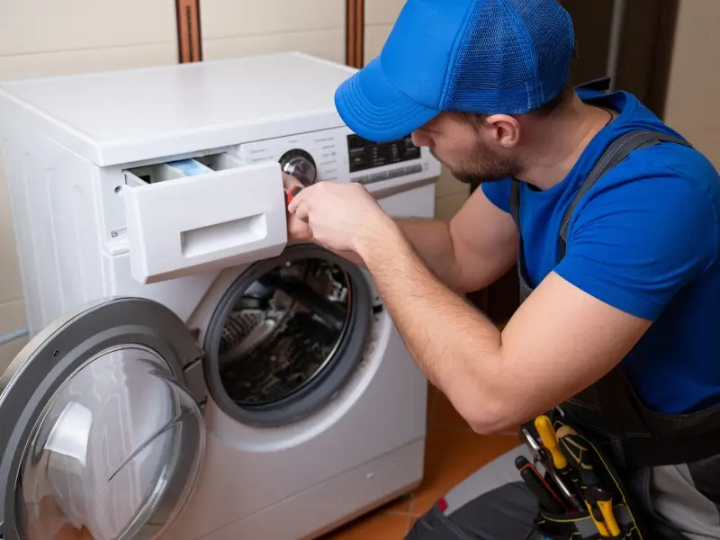 Technician in a blue cap and shirt kneels beside a front-loading washing machine, reaching into the open detergent drawer while holding the machine’s front panel area.