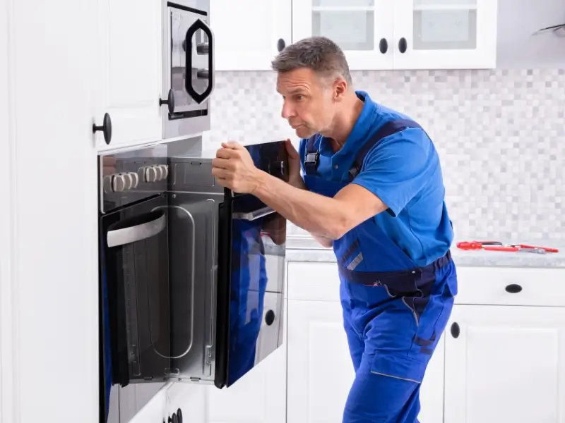A technician in blue workwear completes a wall oven installation in a bright white kitchen, adjusting appliance while standing in front of white cabinets and tiled backsplash.