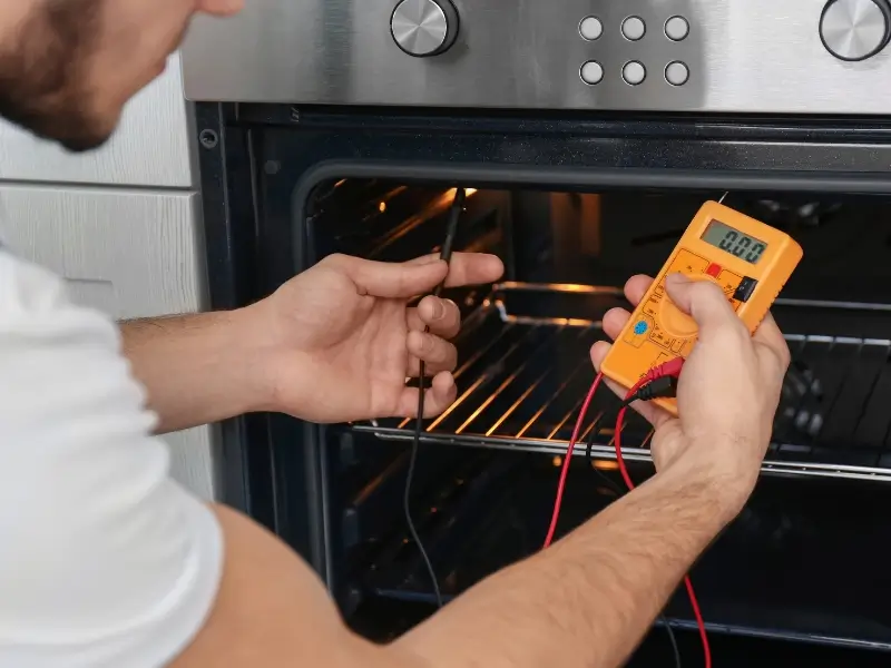 Wall oven repair Technician is holding a metal probe and an orange multimeter inside an open oven, with wires connected to a heating element while the oven interior glows.