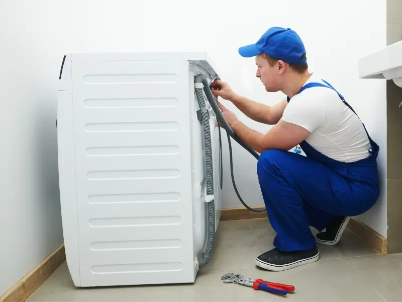 Appliance service technician in a blue cap does a washer dryer installation, working on the washing machine with tools on the floor nearby.