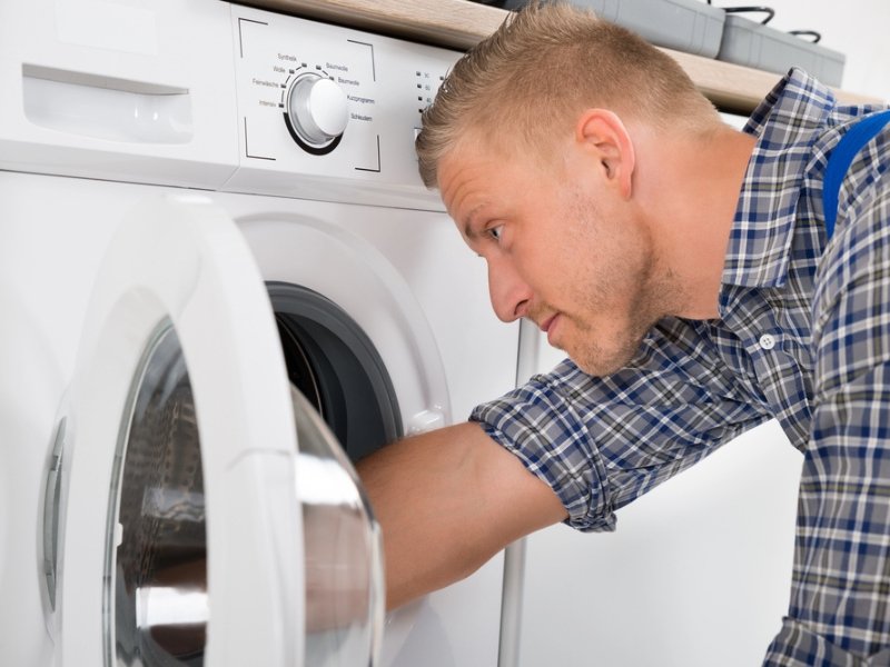 A person in a blue plaid shirt leans over a front-loading washing machine, inspecting and adjusting the control panel and door area.